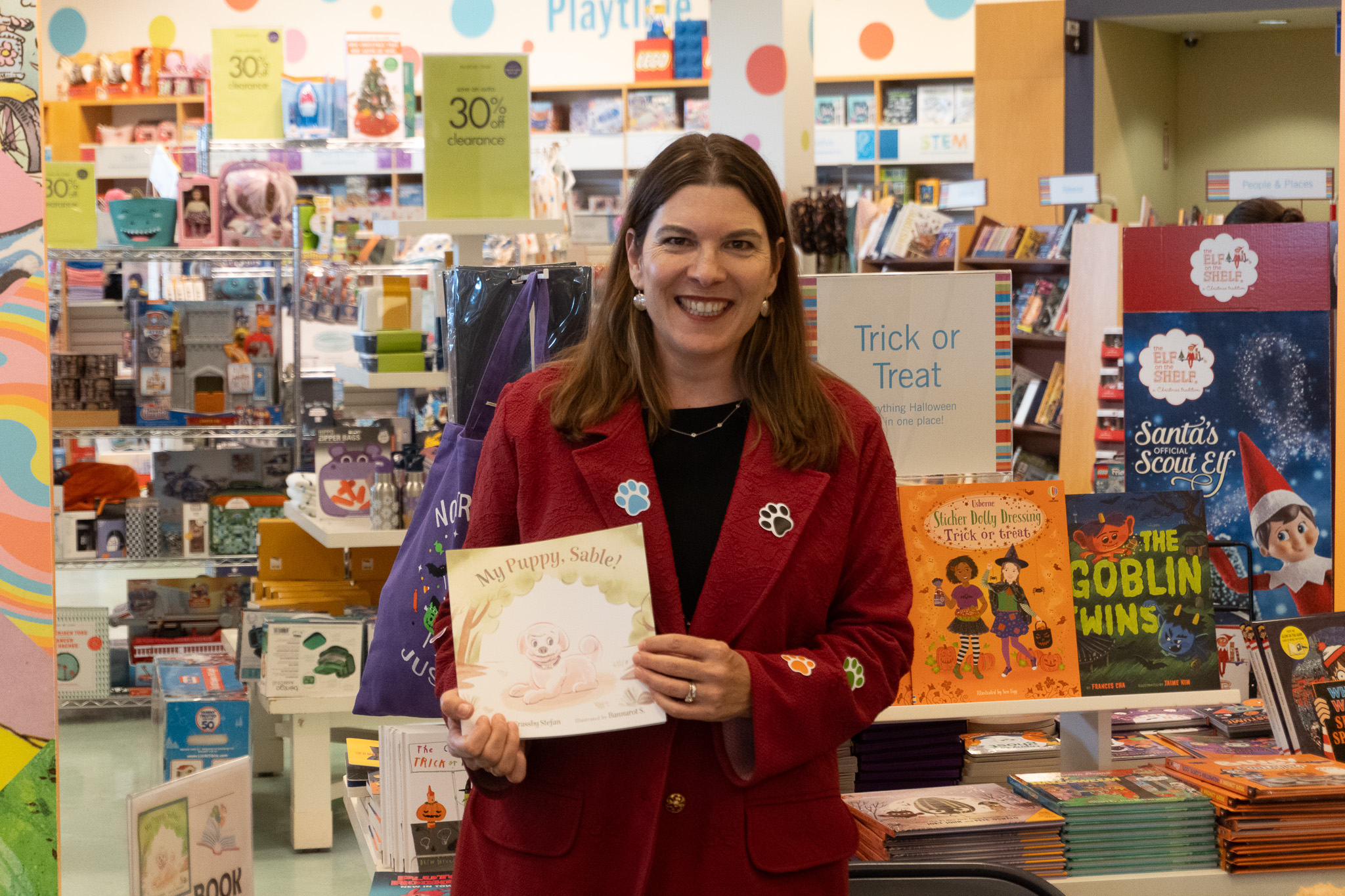 Liz, in a red blazer covered with coloured paw prints, holds up a copy of My Puppy Sable!