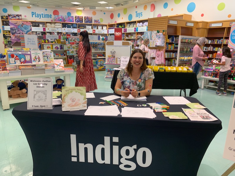 Liz is sitting at a table in Indigo, signing copies of My Puppy Sable! with coloured pencils.